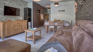 Un salon moderne avec un grand canapé marron, des meubles en bois, un téléviseur fixé à un mur en pierre et un coin repas comprenant une table et des chaises.