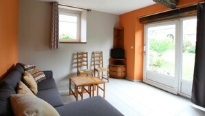 A living room with a sofa, chairs, a small table, a television, and a sliding glass door leading to a garden. - Cozy Stone Farmhouse with Garden, Near Saint-Lô – Sleeps 4, Modern Amenities, Pet-Friendly (Baudre)