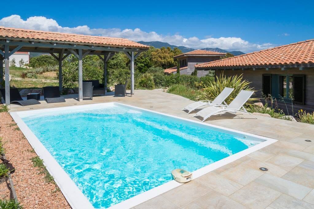 Una piscina rectangular con una pérgola y sillas de descanso, rodeada por un patio con suelo de baldosas.