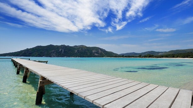 A wooden pier extending into clear turquoise waters, leading to a beach with distant green hills under a partly cloudy sky. - Beautiful villa ' Les Figuiers' with pool between sea and mountain (Figari)