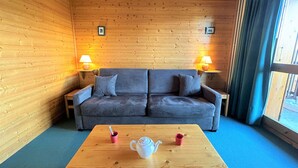 A cozy room with a wooden floor, a grey sofa, two side tables with lamps, a wooden coffee table, and a window with curtains. - 2-room apt for 4, 150m from slopes, Méribel (Méribel)