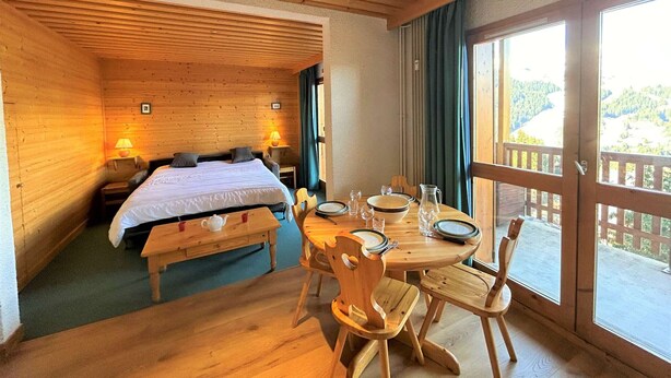 A wooden cabin room with a bed, a dining table with chairs, and a balcony with a view of the outdoors. - 2-room apt for 4, 150m from slopes, Méribel (Méribel)