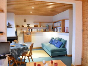 Een gezellige woonkamer met een blauwgroene bank, een houten tafel met een tafelkleed met stippen, en een boekenplank vol boeken.