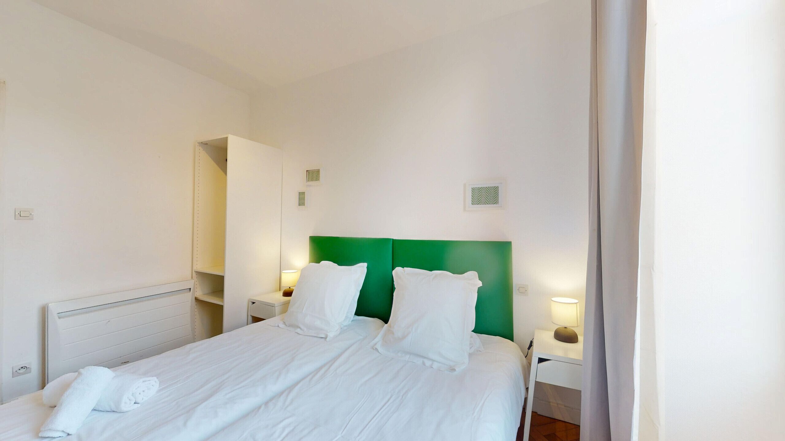 Une chambre d’hôtel avec un grand lit, du linge de lit blanc, une tête de lit verte, deux lampes de chevet, une table de chevet et un miroir.