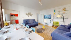 Ein modernes Wohnzimmer mit einem blauen Sofa, einem Esstisch gedeckt mit Tellern und Gläsern, einer Küchenzeile mit einem roten Schrank und einem an der Wand befestigten Fernseher.
