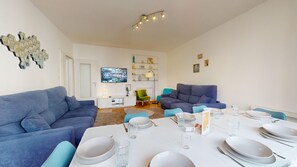 A modern living room with two blue sofas, a dining table set with plates and glasses, and a flat-screen TV mounted on the wall.