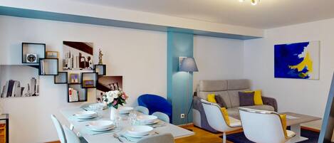 Ein modernes Esszimmer mit Tisch für vier Personen, einem blauen Sessel, einem grauen Sofa mit gelben Kissen und einem Stadtbild an der Wand.
