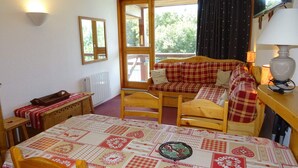 Una habitación acogedora con una cama de madera, una mesa con mantel estampado, una silla, una mesita con bandeja y una ventana con cortinas.