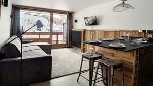 A modern kitchen with a wooden ceiling, a large window with a ski slope view, a flat-screen TV, and a bar area with stools.