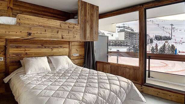 A bedroom with a wooden headboard, a large bed with a quilted duvet, and a window offering a view of a ski slope.