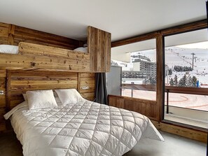 A bedroom with a wooden headboard, a large bed with a quilted duvet, and a window offering a view of a ski slope. - Renovated 36m² apartment, ski-in/out, south-facing balcony (Les Menuires)