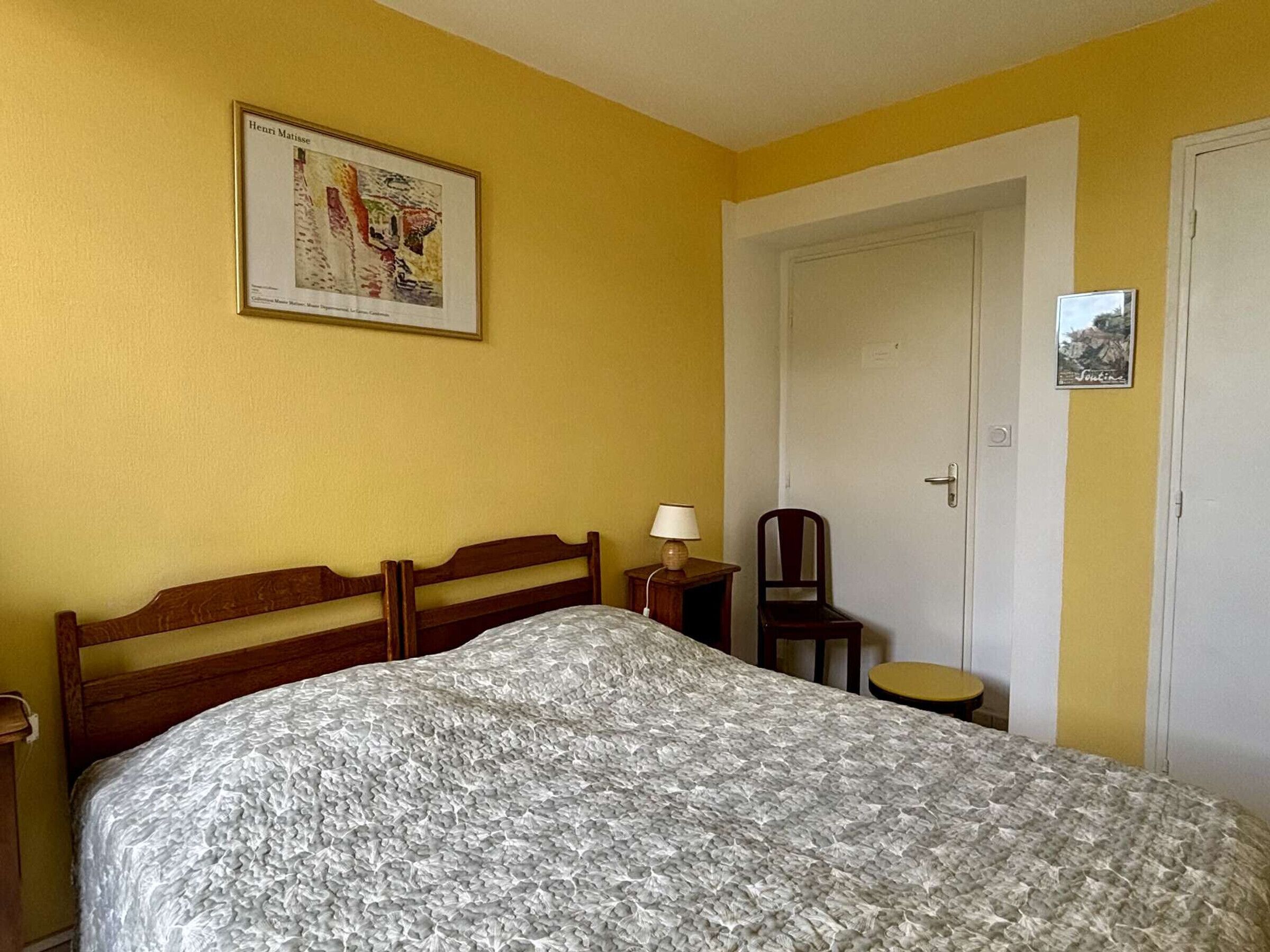 Un dormitorio con cama, silla, mesita de noche, puerta y un cuadro en la pared.