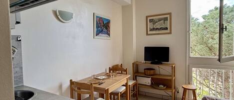 A compact living space with a dining area, a TV on a shelf, and a window with a view of trees.
