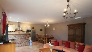 Una sala de estar con un sofá, una mesa de centro de madera, una mesa de comedor con sillas, una cocina con armarios y electrodomésticos, y un candelabro.