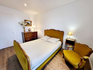 A neatly arranged bedroom with a bed, a wooden dresser, a chair, and a nightstand with a lamp.