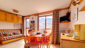 A cozy room with a wooden sofa, a dining table set with plates and glasses, a coffee maker, and a window with a view of mountains. - Functional studio at the foot of the slopes with balcony (Les Menuires)