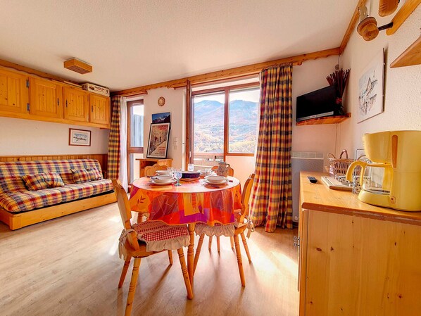 Ein gemütliches Zimmer mit einer Holzdirektiva, einem Esstisch gedeckt mit Tellern und Gläsern, einer Kaffeemaschine und einem Fenster mit Bergblick.