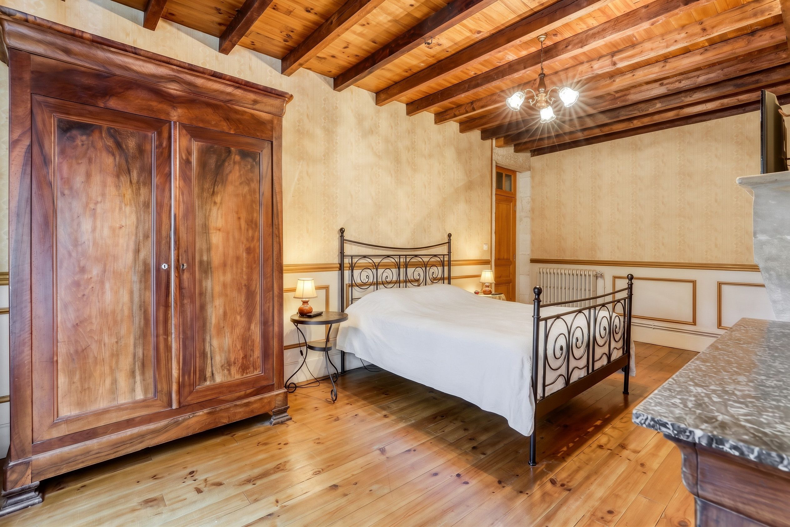 A bedroom with a wooden floor, a bed with a metal frame, a wooden wardrobe, and a small bedside table with a lamp.