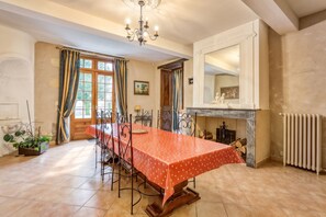 A dining area with a long table, chairs, a chandelier, a fireplace, and a window with curtains. - Homerez - Spacious House - Marcellus - 400 m² - Garden (Marcellus)