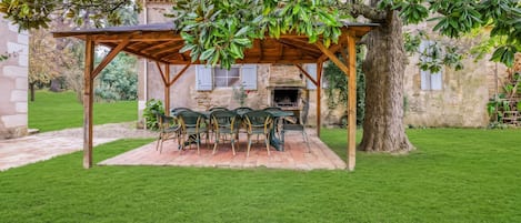 A covered outdoor seating area with a stone fireplace, surrounded by a well-maintained lawn and trees.