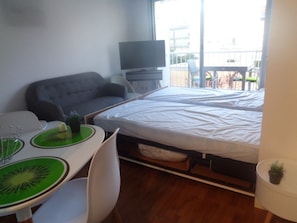 Um espaço de estar compacto com um sofá, uma cama, uma televisão, uma mesa de jantar com um jogo americano verde e uma pequena planta em vaso.