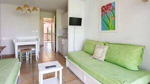 A room with a green sofa, a small table, a TV mounted on the wall, and a painting of flowers.