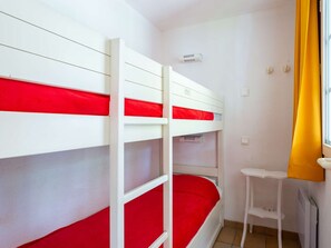 Een slaapkamer met een stapelbed, een rode matras, een klein wit tafeltje en een raam met gele gordijnen.