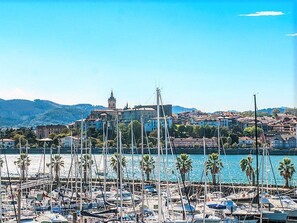 A marina with numerous boats docked, a prominent building with a tower in the background, and a hilly landscape.