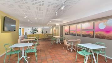 A modern dining area with green chairs and white tables, a flat-screen TV on the wall, and a sunset view through the windows.