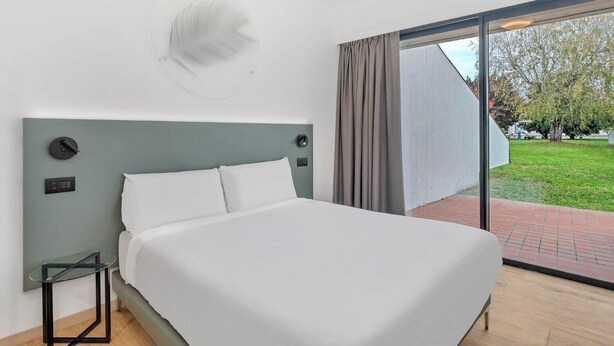A modern bedroom with a large bed, a glass table, and a view of a garden through a sliding glass door.