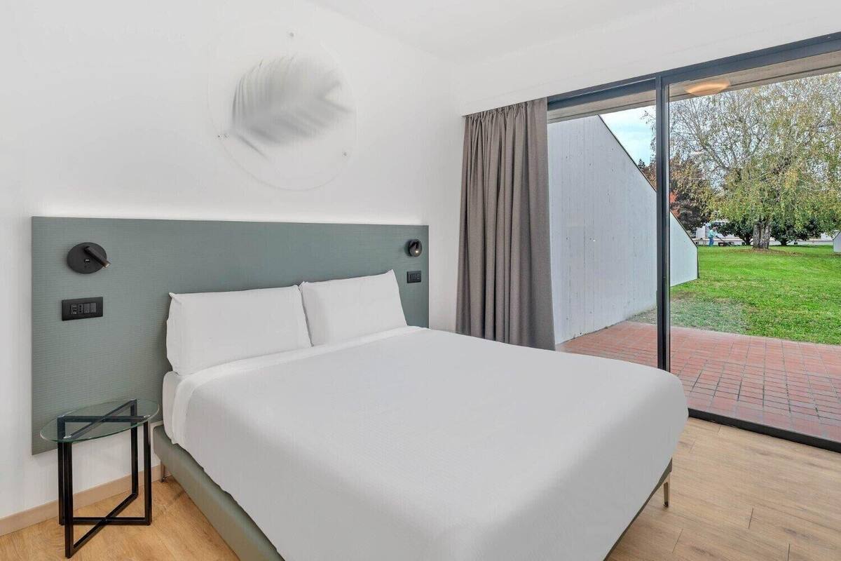 Une chambre moderne avec un grand lit, une table en verre et une vue sur un jardin, accessible par une porte-fenêtre coulissante.
