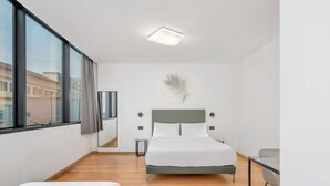 A modern hotel room with a large bed, a desk, and a view of the city through large windows. - B&B Hotel Alba (Alba)