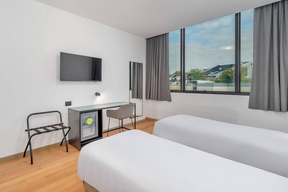 Une chambre d’hôtel moderne dotée d’une grande fenêtre, d’un bureau avec une chaise, d’un lit et d’un téléviseur à écran plat fixé au mur.