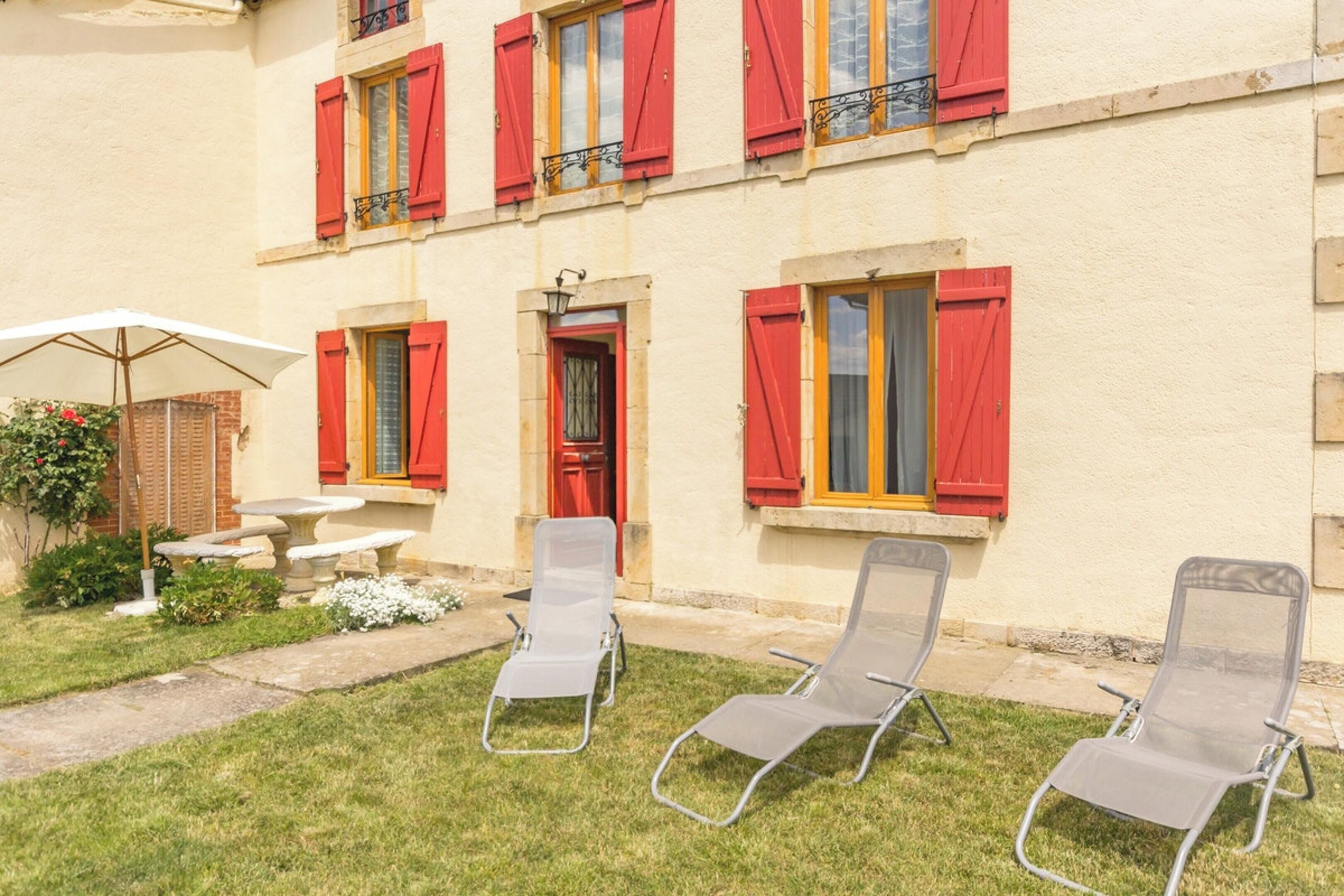 A building with red shutters, a stone bench, and two lounge chairs in the garden.