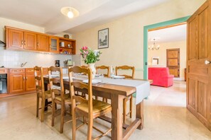 Un coin repas avec des chaises en bois et une table dressée pour quatre personnes, une cuisine avec des meubles en bois et un dosseret carrelé, et une chaise rouge en arrière-plan.