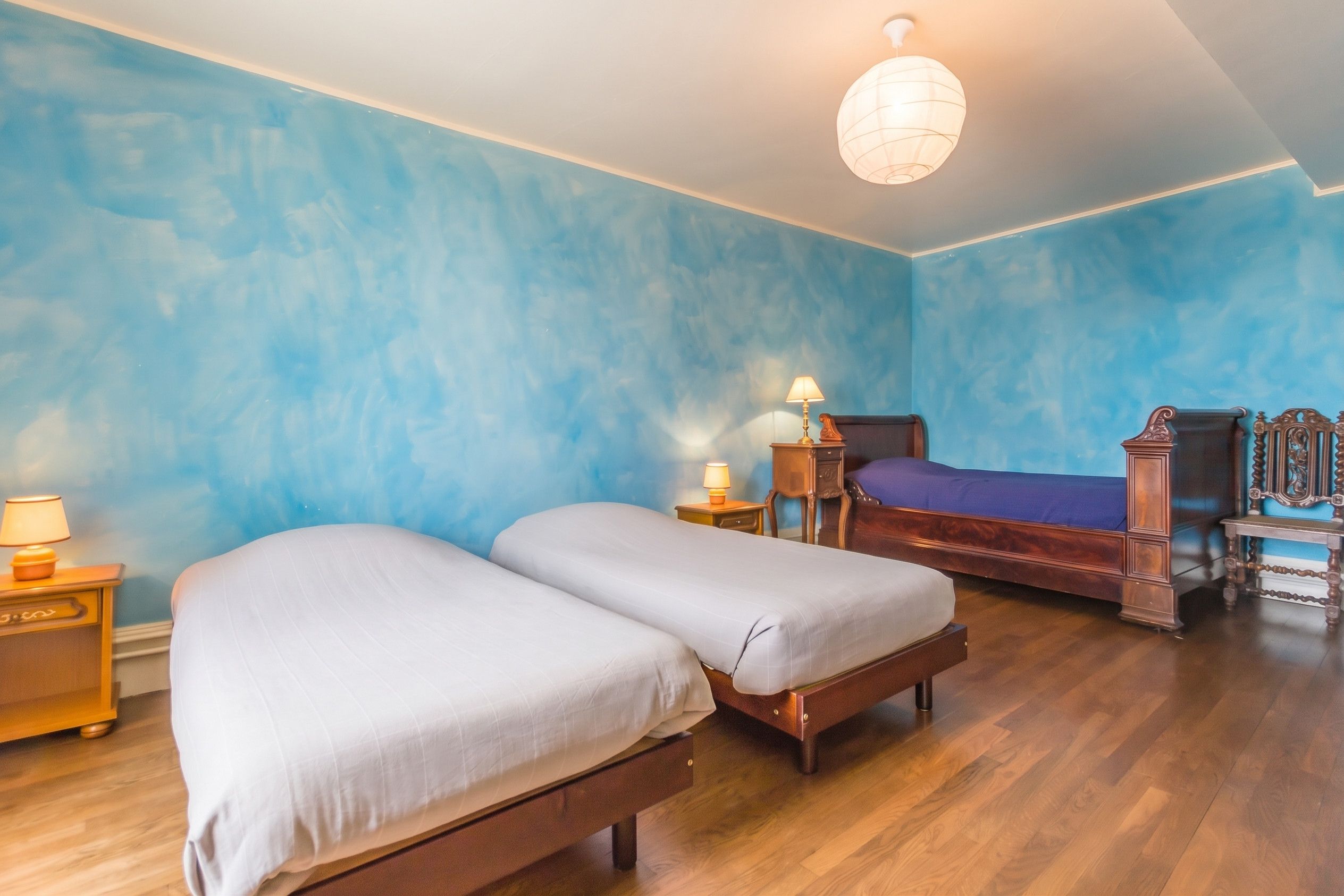 Une chambre avec deux lits, un plancher en bois, un mur bleu et un luminaire suspendu.