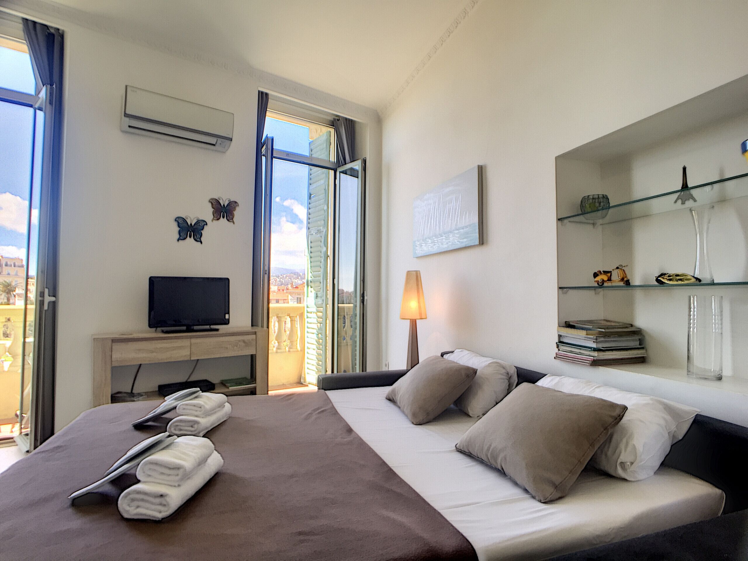 A hotel room with a bed, a television, a desk, a lamp, and a view of the city.