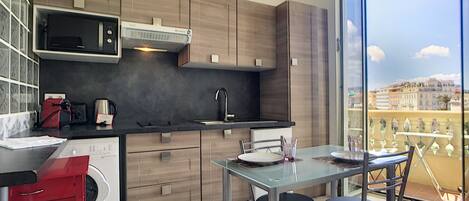 A modern kitchen with a built-in microwave, a sink, and wooden cabinets. There is a dining table with two chairs and a view of the city through large windows.