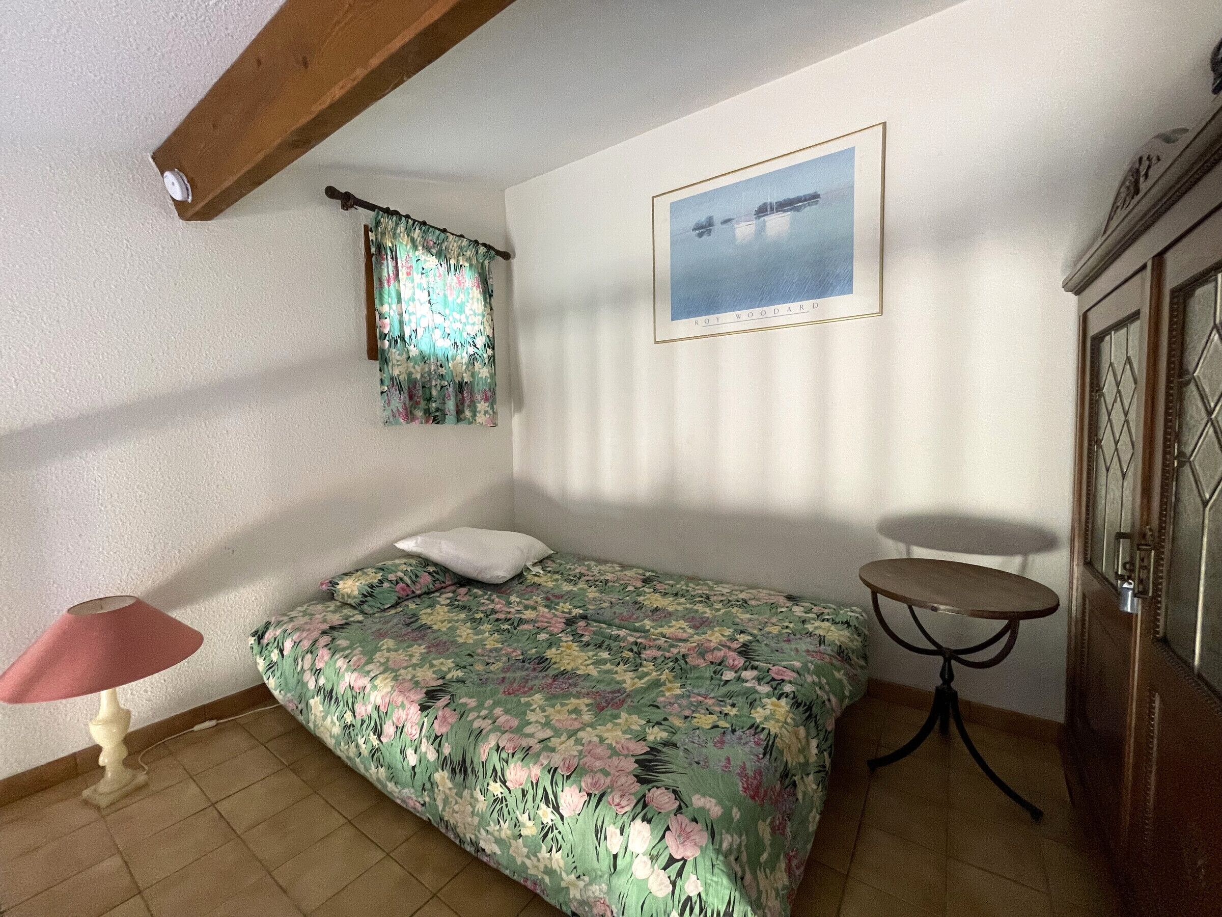 Ein Schlafzimmer mit einer blumigen Bettdecke, einem weißen Kopfkissen, einem kleinen runden Tisch und einem gerahmten Bild an der Wand.