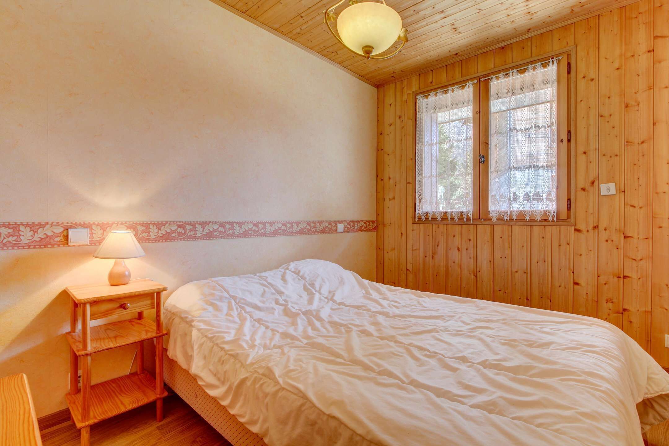 Un dormitorio con techo de madera, una cama con ropa de cama blanca, una mesita de noche con lámpara y una ventana con cortinas de encaje.