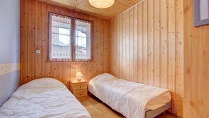 A small, cozy room with wooden walls and floors, two single beds, a nightstand with a lamp, and a window with lace curtains. - Sermes 1 - Spacious apartment in the centre (MORZINE)