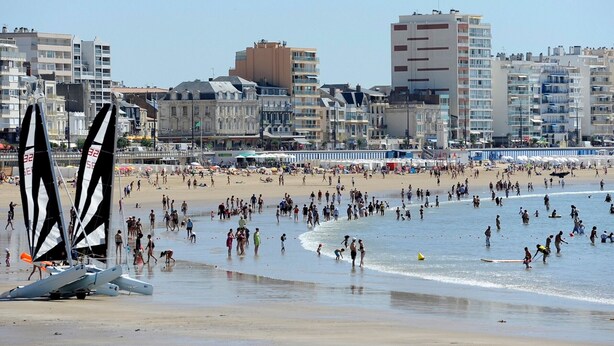 Une plage avec des gens, un voilier et des bâtiments en arrière-plan.
