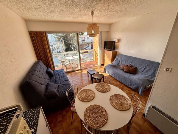 Ein kompakter Wohnraum mit einer Couch, einem kleinen Tisch mit gewebten Untersetzern, einem Fernseher und einem Balkon mit Gartenmöbeln.