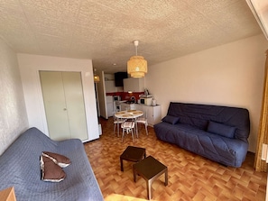 Un espace de vie compact comprenant un canapé, une table à manger avec des chaises, un coin cuisine et un plafond orné d’un motif texturé.