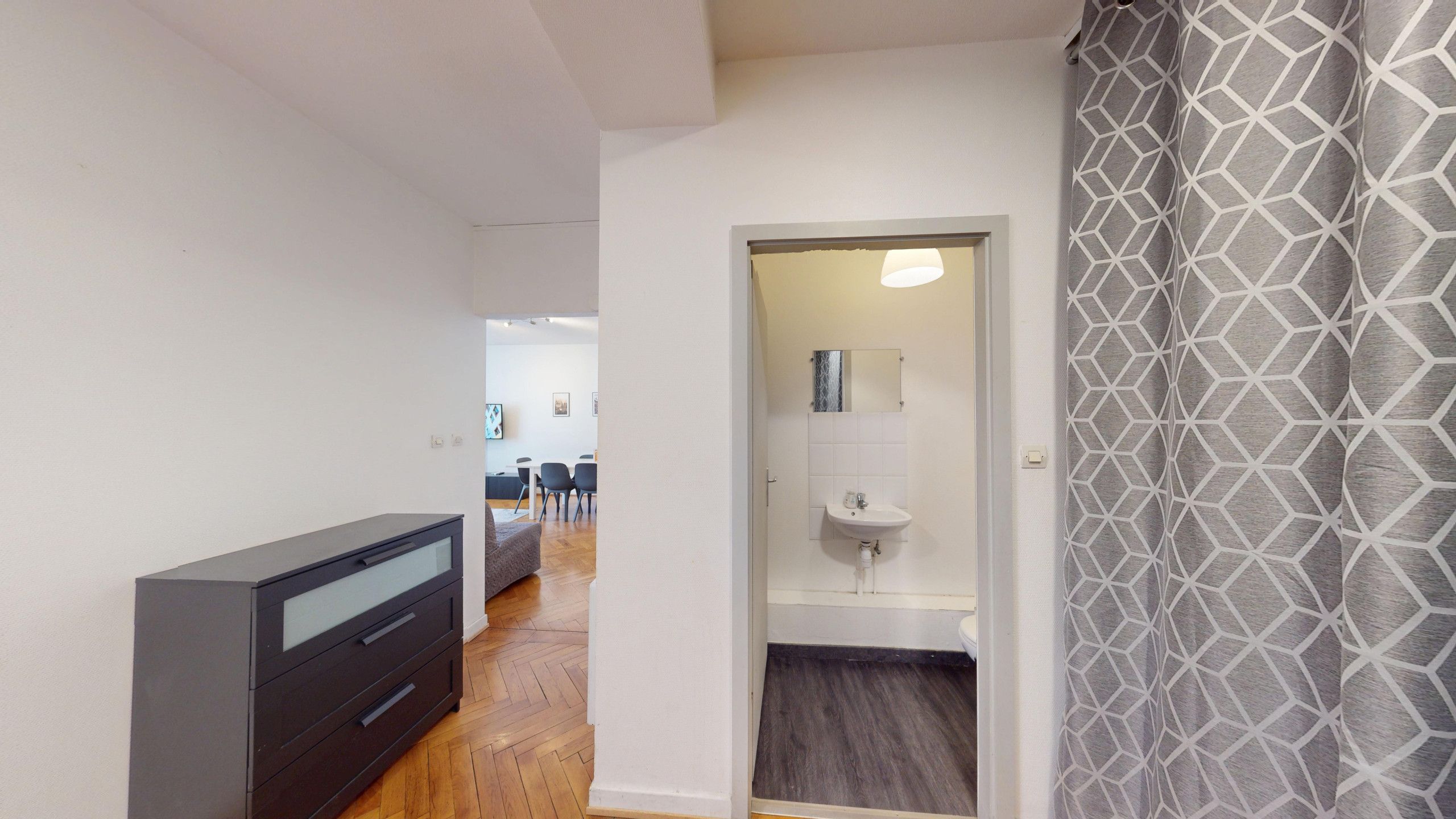 Un couloir avec un meuble de rangement de couleur sombre, une salle de bain avec un lavabo et un miroir, et un rideau à motifs.