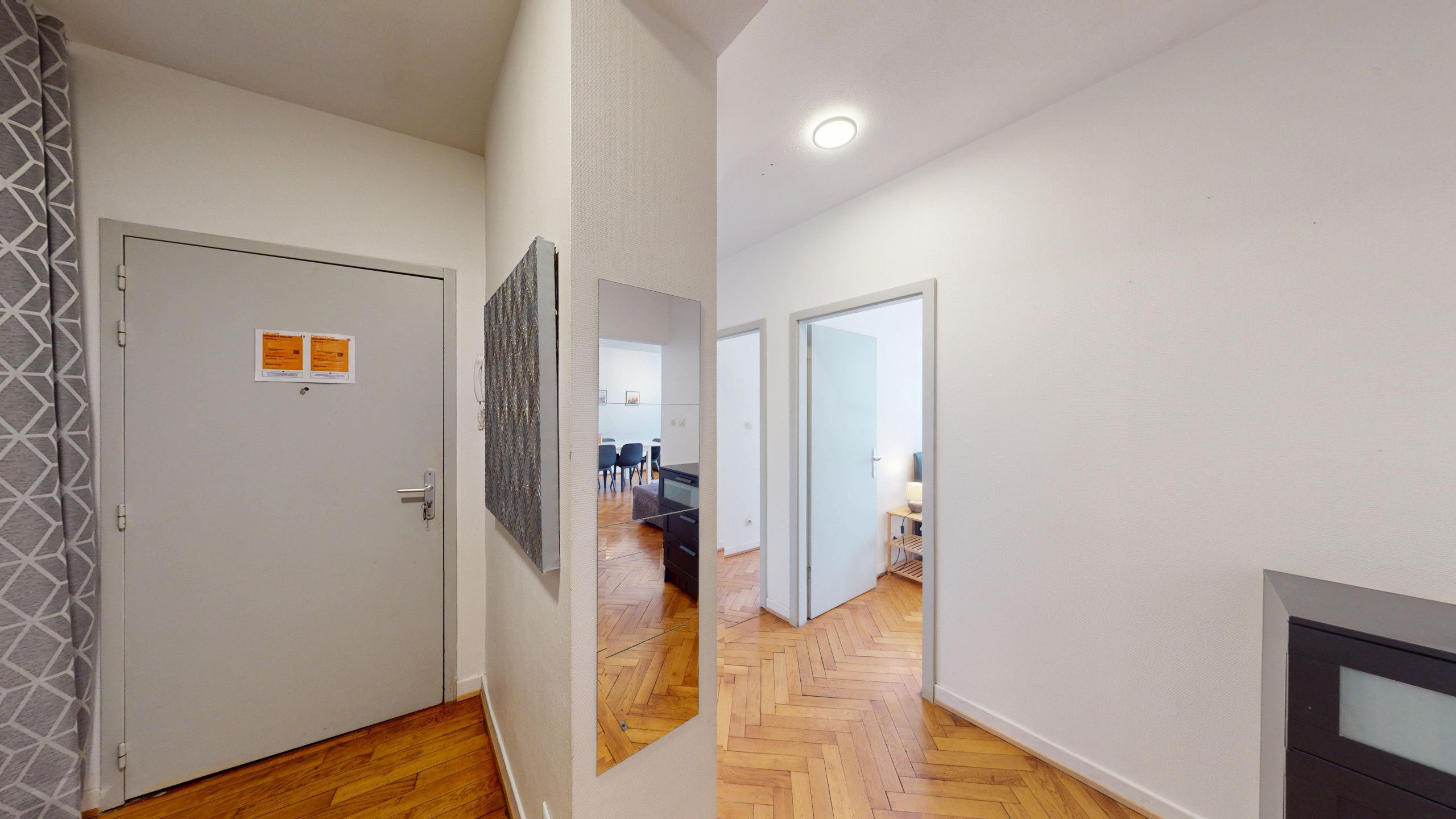 Un couloir avec une porte, un panneau fixé au mur, et une vue sur une pièce où l’on aperçoit un canapé et une table.