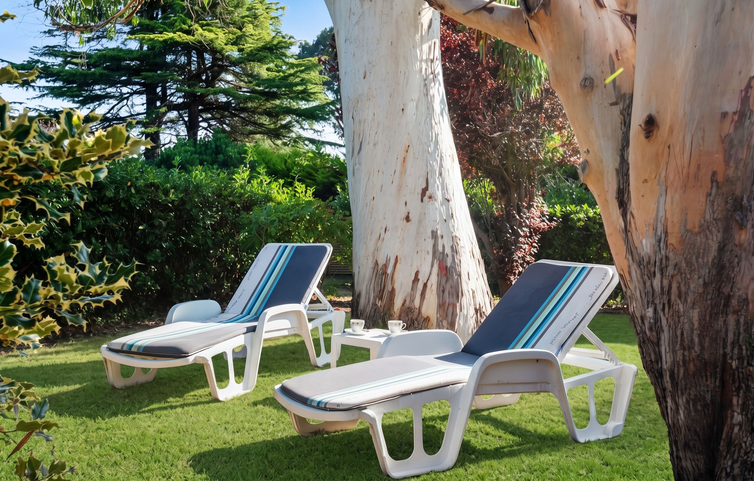 Two lounge chairs with striped cushions, a white table, and a cup of tea, set on a grassy lawn with trees in the background.