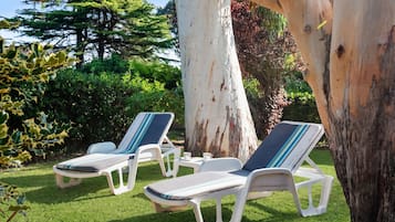 Two lounge chairs with striped cushions, a white table, and a cup of tea, set on a grassy lawn with trees in the background.