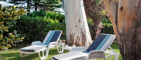 Two lounge chairs with striped cushions, a white table, and a cup of tea, set on a grassy lawn with trees in the background.