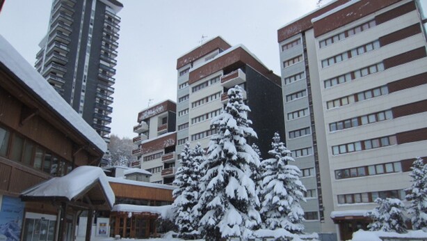 A snow-covered courtyard with a building featuring multiple floors and balconies, surrounded by snow-laden trees. - Spacious ski-in/ski-out apartment with balcony (Les Menuires)